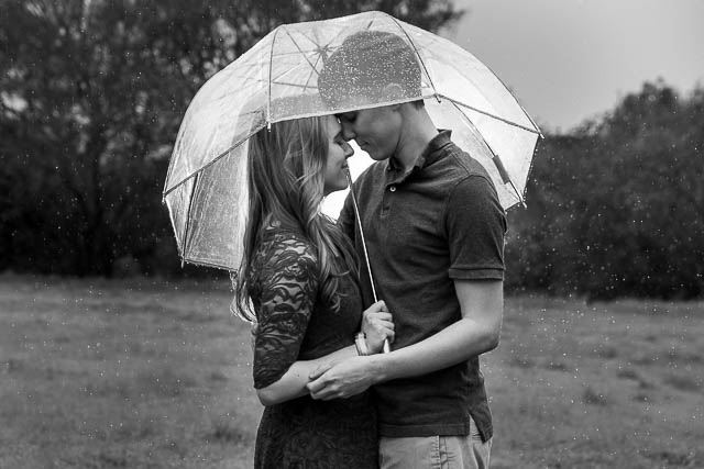 Engaged couple holding each other under the clear umbrella in the rain at Hans Christian Andersen Park in Solvang California.