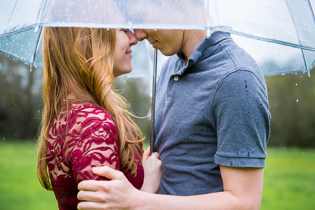 Engaged couple holding each other under the clear umbrella in the rain at Hans Christian Andersen Park in Solvang California.