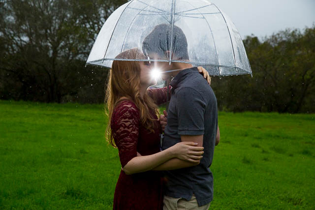Engaged couple holding each other under the clear umbrella in the rain at Hans Christian Andersen Park in Solvang California.