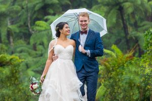 Bride and groom using a clear umbrella to shield them from the rain during their Green Lake wedding photos in Rotorua, New Zealand.